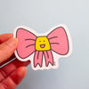 A hand holds a die-cut magnet featuring a pink bow with a yellow smiling face in the center, against a light blue background.