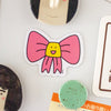 A die-cut magnet featuring a pink bow with a yellow smiling face in the center, shown on a light surface with other decorative items partially visible around it.