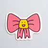 A die-cut magnet featuring a pink bow with a yellow smiling face in the center, shown on a light blue background.