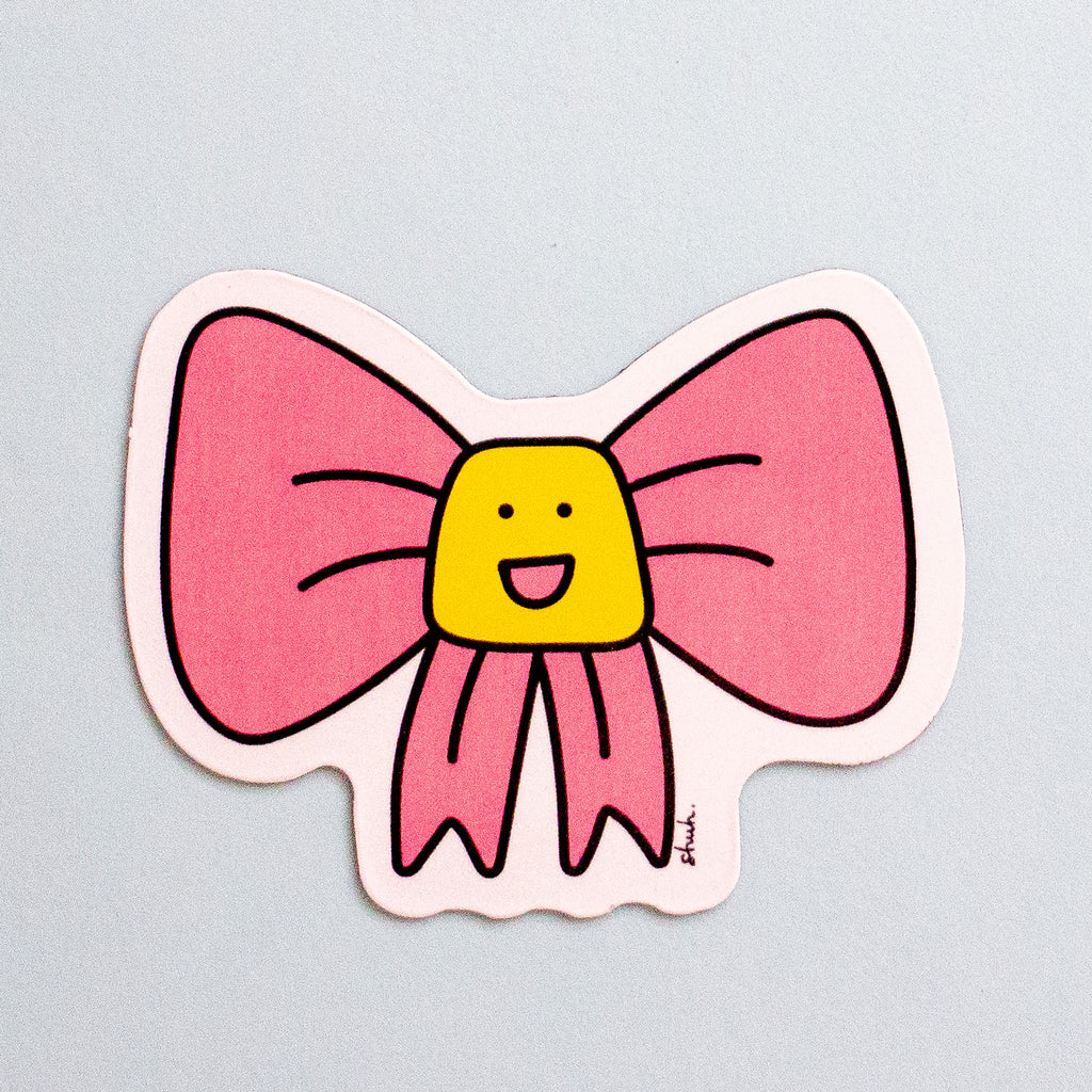 A die-cut magnet featuring a pink bow with a yellow smiling face in the center, shown on a light blue background.
