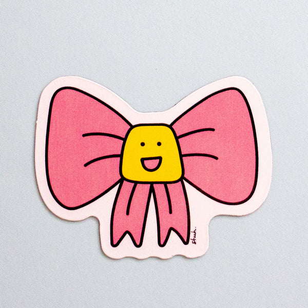A die-cut magnet featuring a pink bow with a yellow smiling face in the center, shown on a light blue background.