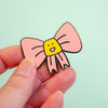 A hand holding a small enamel pin shaped like a pink bow with a yellow smiling face in the center, against a pale mint background.