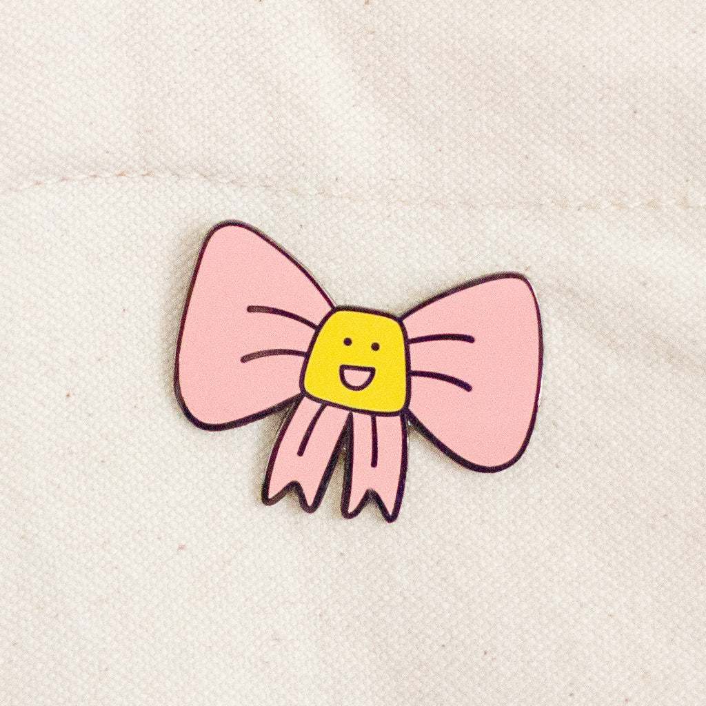 A small enamel pin shaped like a pink bow with a yellow smiling face in the center, placed on a light beige fabric background.