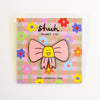 A pink bow enamel pin with a yellow smiling face in the center, mounted on a decorative pink and beige gingham backing card with colourful flowers and the brand name 'shuh.'.