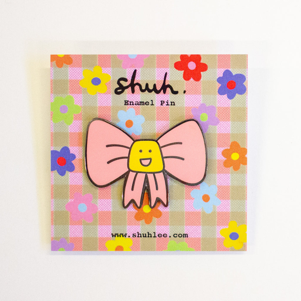 A pink bow enamel pin with a yellow smiling face in the center, mounted on a decorative pink and beige gingham backing card with colourful flowers and the brand name 'shuh.'.