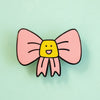 A pink bow enamel pin with a yellow smiling face in the center, set against a solid mint green background.
