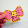 A hand holding a die-cut sticker featuring a cute pink bow cartoon character , set against a plain light grey background.