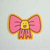 A die-cut sticker featuring a cute pink bow cartoon character , set against a plain light grey background.