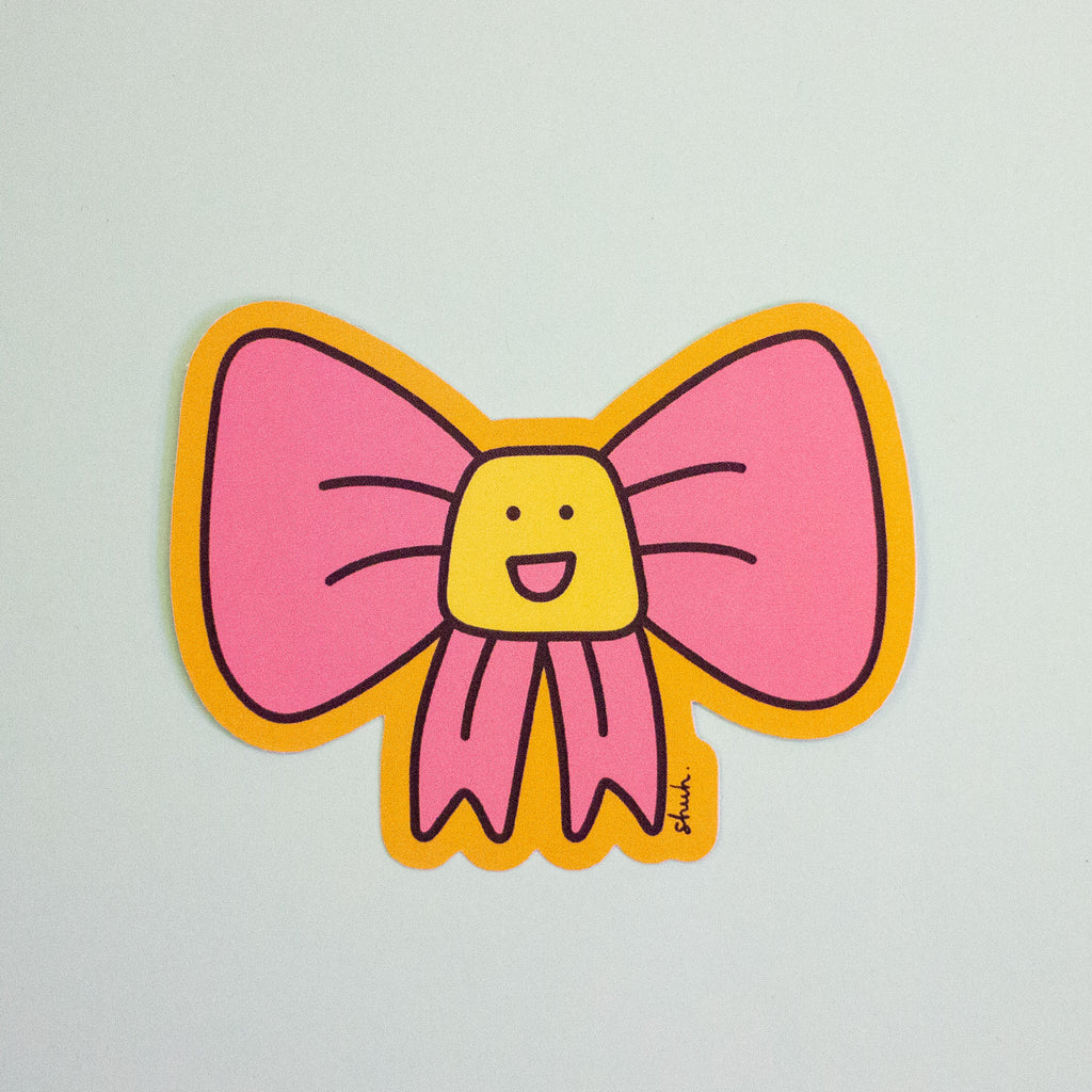 A die-cut sticker featuring a cute pink bow cartoon character , set against a plain light grey background.