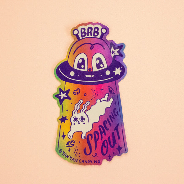 A holographic sticker featuring a spaceship and a cartoon illustration with 'BRB Spacing Out' text. 