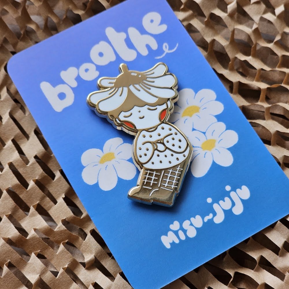 A cute enamel pin of a girl with a flower crown sitting among daisies, mounted on a blue backing card that reads 'breathe' and 'Misu Juju'.
