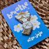 A cute enamel pin of a girl with a flower crown sitting among daisies, mounted on a blue backing card that reads 'breathe' and 'Misu Juju'.