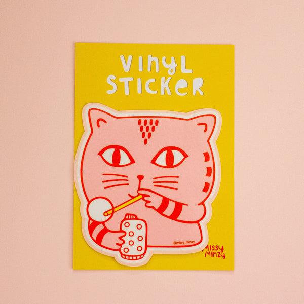 A vinyl sticker featuring a pink cartoon cat holding a straw stick and blowing a bubble on a bright yellow backing card, displayed against a soft pink background.