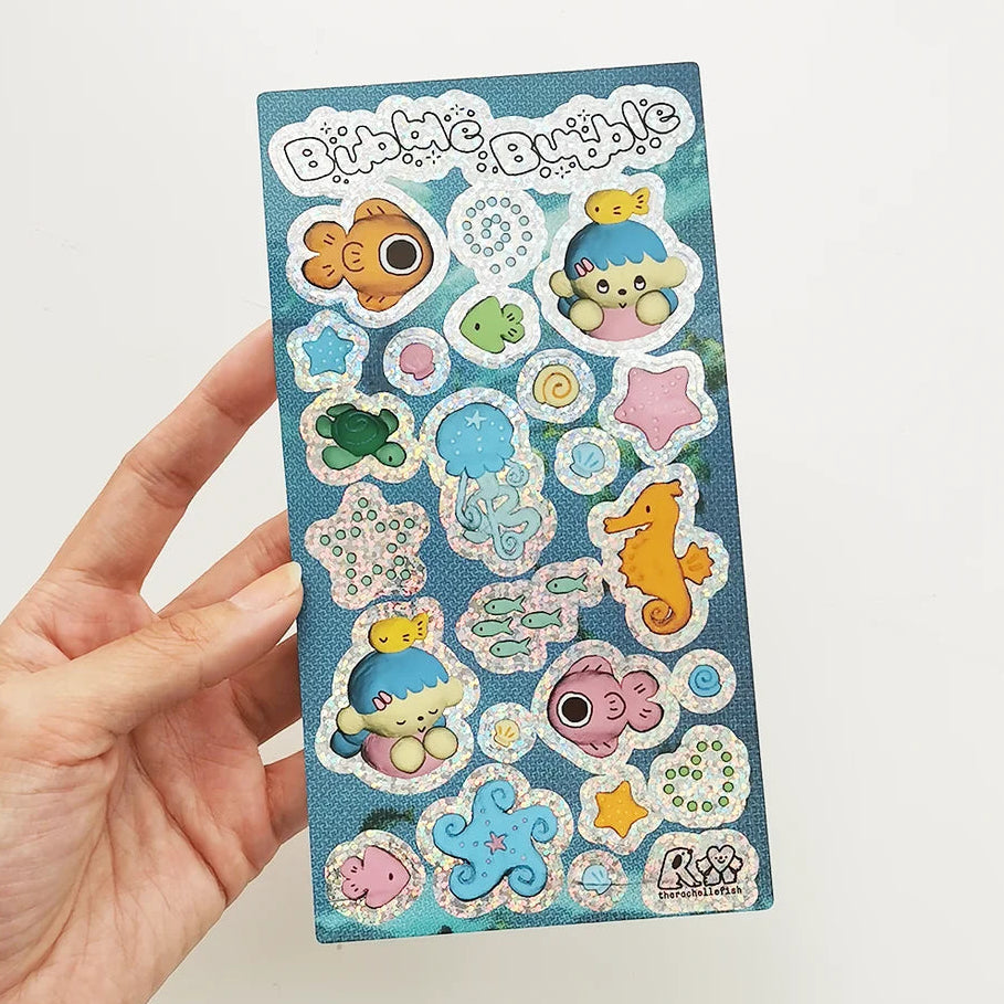 Hand holding a glitter sticker sheet featuring sea-themed illustrations with title 'Bubble Bubble'. 