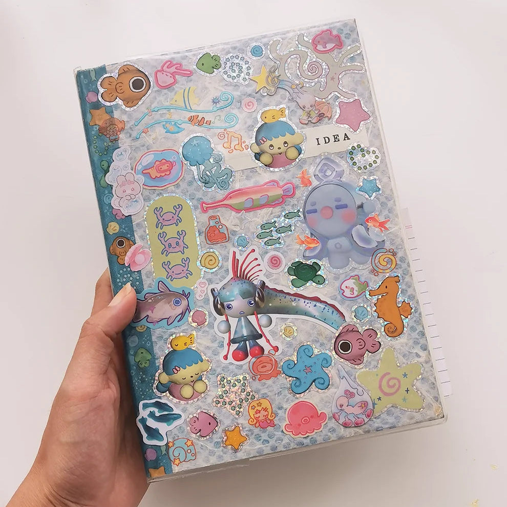 Hand holding a journal featuring a range of stickers including glitter stickers of sea-themed cartoons. 