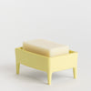 A pale yellow rectangular soap dish with four legs holding a translucent cream-coloured bar of soap, photographed on a clean white studio background.