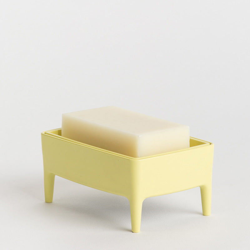 A pale yellow rectangular soap dish with four legs holding a translucent cream-coloured bar of soap, photographed on a clean white studio background.