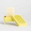 A bright yellow plastic soap dish featuring a rectangular stand with a small leg and a soap, alongside a separate perforated yellow lid or cover, photographed on a clean light grey studio background.