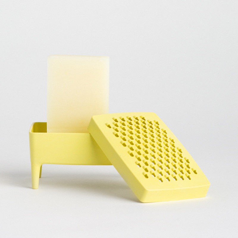 A bright yellow plastic soap dish featuring a rectangular stand with a small leg and a soap, alongside a separate perforated yellow lid or cover, photographed on a clean light grey studio background.