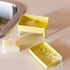 Two small yellow soap dish parts with feet, one holding a bar of soap and the other containing shredded soap, on a beige countertop near a sink.