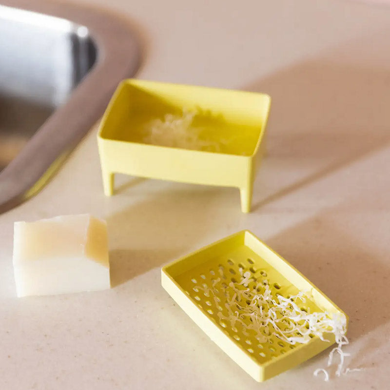 Two small yellow soap dish parts with feet, one holding a bar of soap and the other containing shredded soap, on a beige countertop near a sink.