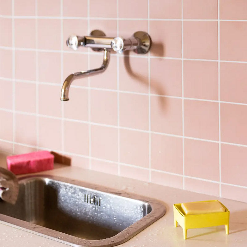 A wall-mounted chrome faucet with clear round knobs installed above a stainless steel sink, set against a pink tiled wall. A pink sponge and a small yellow soap dish sit on the counter.
