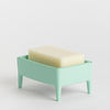 A mint green plastic soap dish with four legs holding a rectangular bar of cream-coloured soap, set against a plain light background.