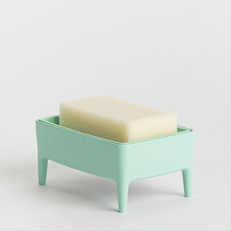 A mint green plastic soap dish with four legs holding a rectangular bar of cream-coloured soap, set against a plain light background.