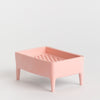A light pink plastic soap dish with four legs and a perforated top tray, photographed on a seamless light grey background.
