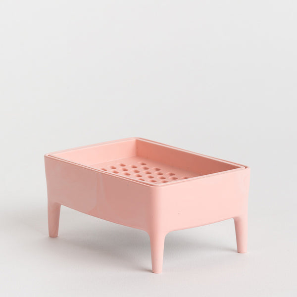 A light pink plastic soap dish with four legs and a perforated top tray, photographed on a seamless light grey background.