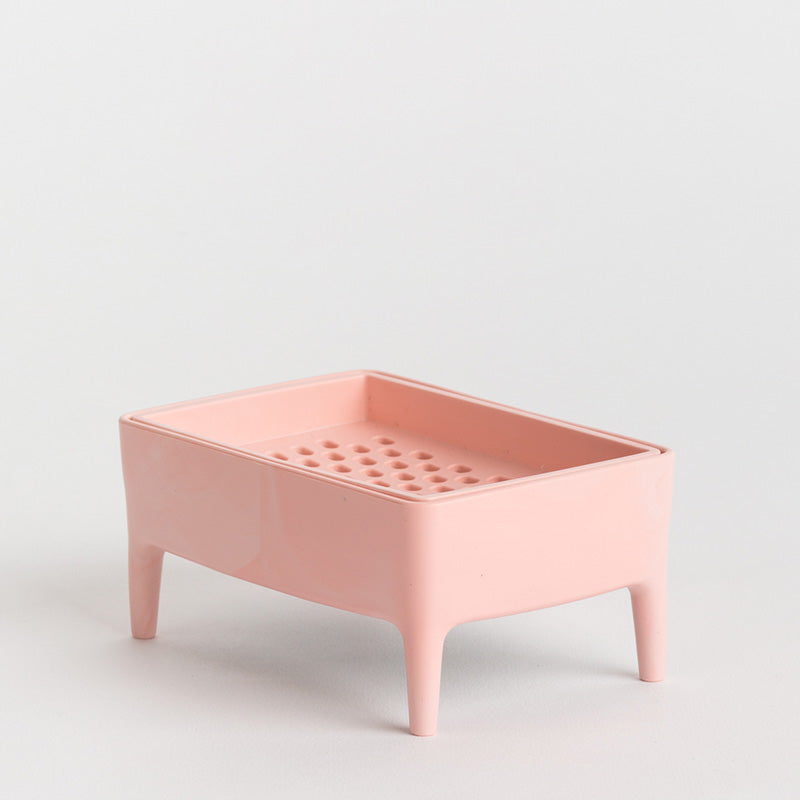 A light pink plastic soap dish with four legs and a perforated top tray, photographed on a seamless light grey background.