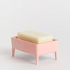 A pink rectangular footed soap dish holding a cream-coloured bar of soap, photographed on a clean white studio background.