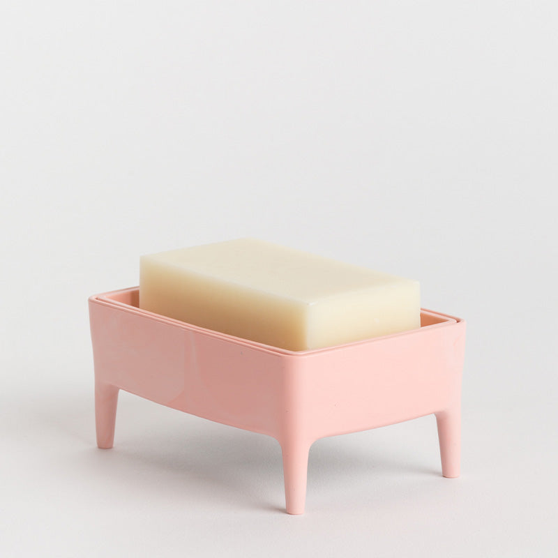 A pink rectangular footed soap dish holding a cream-coloured bar of soap, photographed on a clean white studio background.