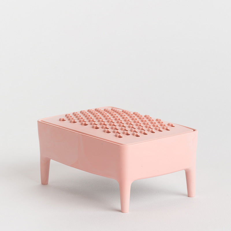 A small pink soap dish with a textured, raised pattern on the top surface and four short legs, photographed on a clean white studio background.