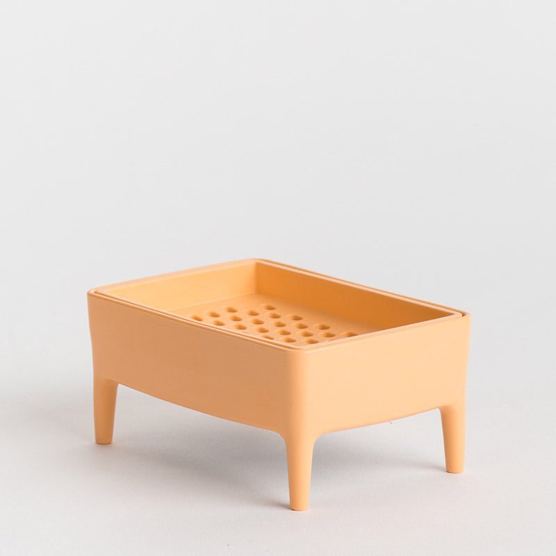 A small rectangular peach-coloured plastic tray with four legs and a perforated top, photographed on a clean light grey background.