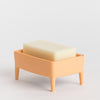 A peach-coloured plastic soap dish with small legs, holding a rectangular bar of creamy off-white soap, set against a plain light grey background.