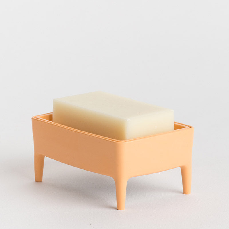 A peach-coloured plastic soap dish with small legs, holding a rectangular bar of creamy off-white soap, set against a plain light grey background.