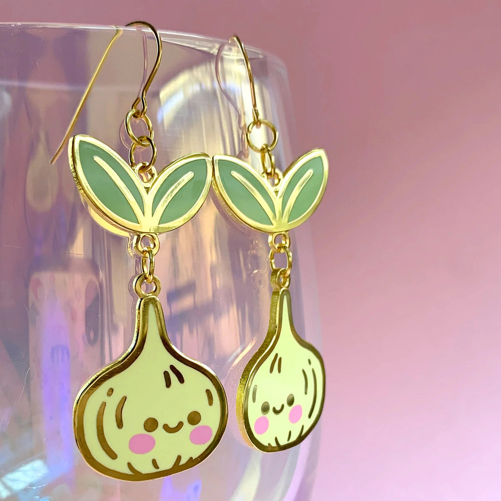 A pair of dangling earrings shaped like smiling green sprouts with gold-tone outlines and hooks, displayed in front of a translucent glass against a soft pink background.