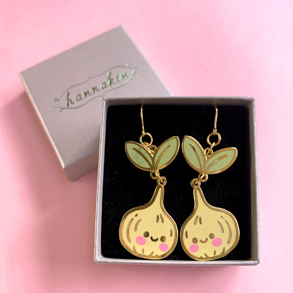 A pair of gold-tone dangle earrings shaped like cute bulb characters with mint green leaves and pink cheeks, displayed in a black-lined gift box next to a silver box lid with the brand name 'hannakin' on a pink background.