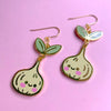 A pair of gold-tone dangle earrings shaped like cute, smiling green onions with pastel green leaves, cream-coloured bodies, and pink cheeks, displayed on a solid pink background.