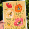 Print with colourful flowers and bees on a yellow background, held against a natural setting.