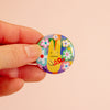 A hand holding a round pinback button featuring a colourful background with a bunny illustration with flowers.