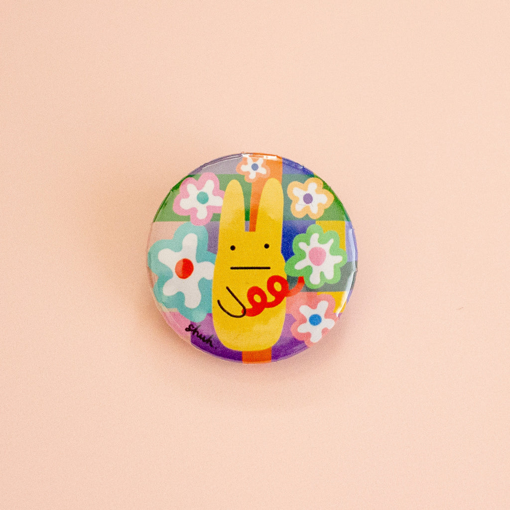 A round pinback button featuring a colourful patchwork background with a yellow rabbit character, displayed on a solid pale peach background.
