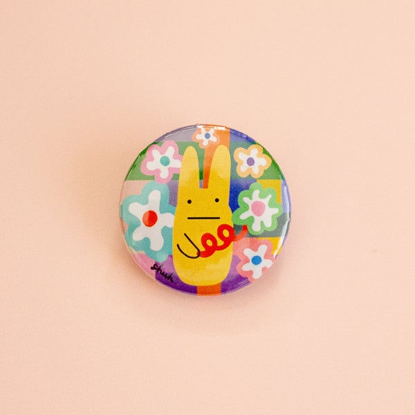 A round pinback button featuring a colourful patchwork background with a yellow rabbit character, displayed on a solid pale peach background.