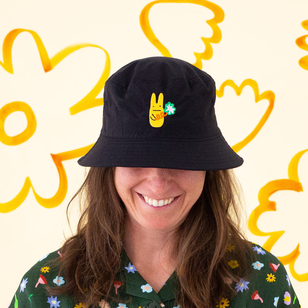 Smiling person wearing a black bucket hat with a yellow bunny and green clover embroidery, paired with a dark green floral shirt, standing in front of a pale yellow background with bold orange flower illustrations