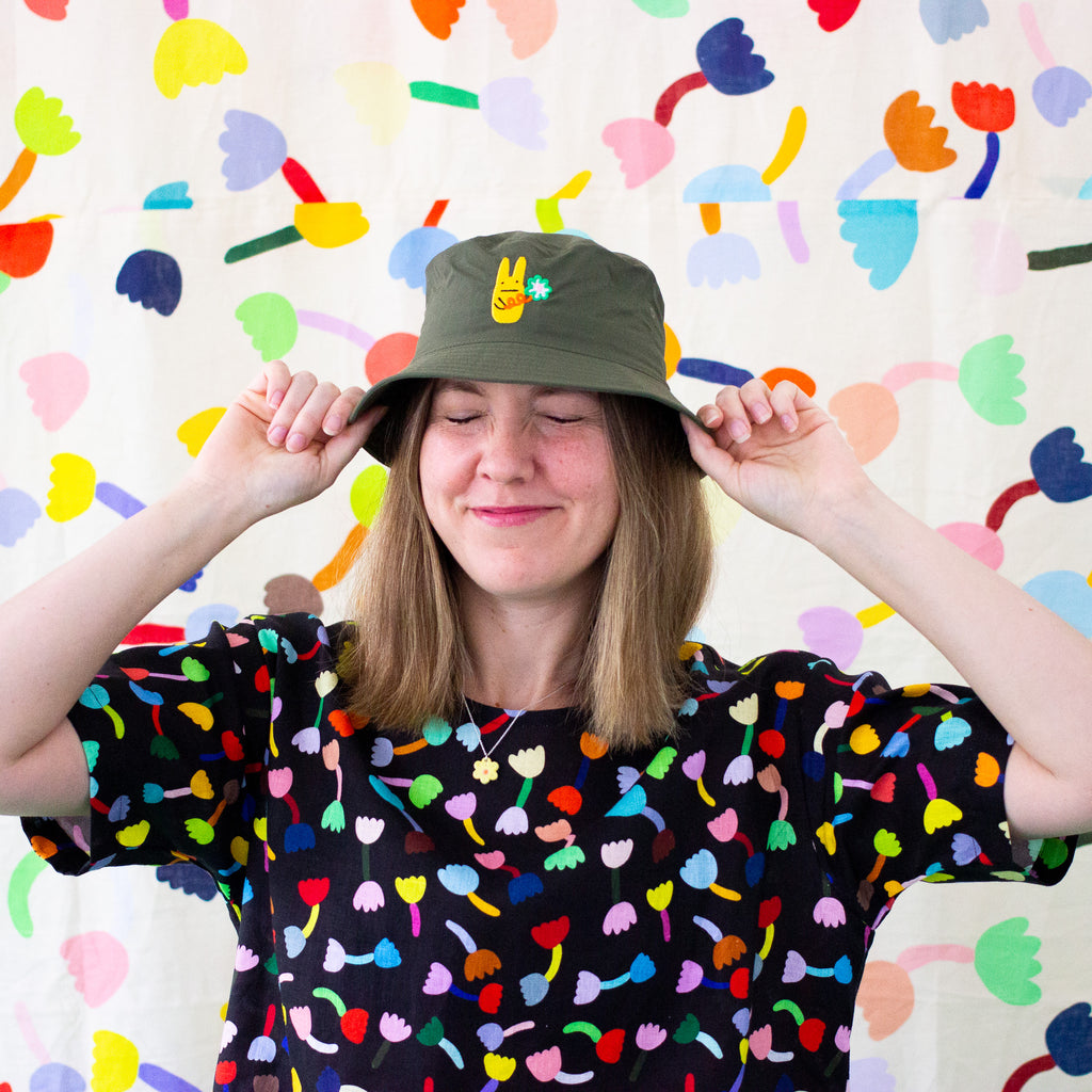 Front view of a model placing the green bunny bucket hat on their head. Add a touch of fun to your outfit with this playful bucket hat, featuring an embroidered yellow bunny character holding a large flower.
