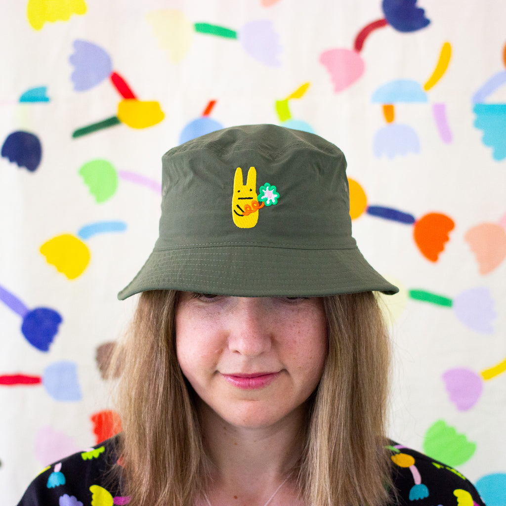 Front view of a model wearing the green bunny bucket hat. Add a touch of fun to your outfit with this playful bucket hat, featuring an embroidered yellow bunny character holding a large flower.