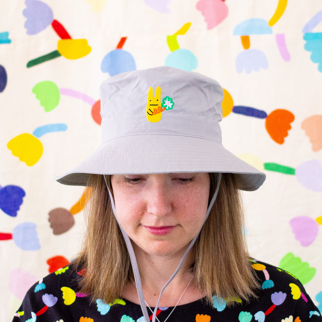 Model wearing the grey bunny bucket hat. Embroidered yellow bunny holding a green flower. 