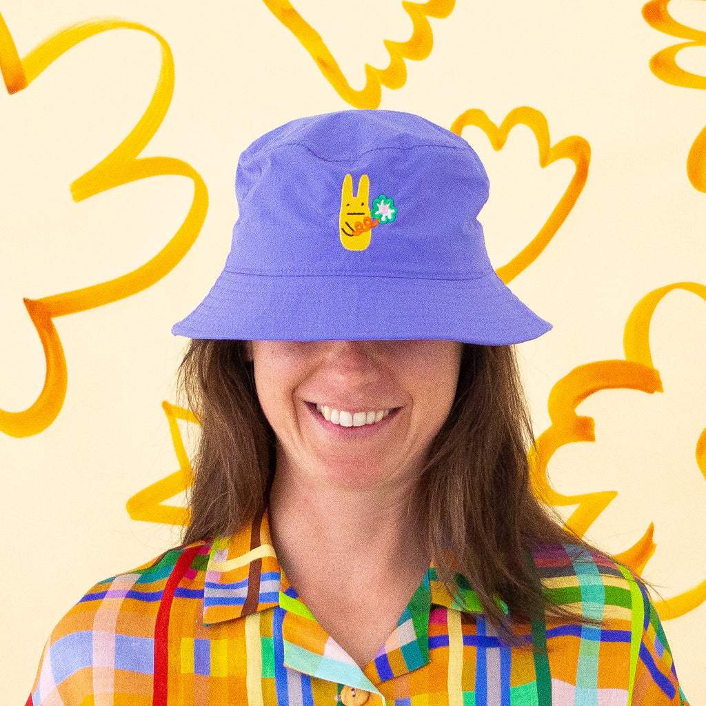 Smiling person wearing a lapis coloured bucket hat with a yellow bunny and green clover embroidery, paired with a colourful checkered shirt, standing in front of a yellow background with abstract orange shapes