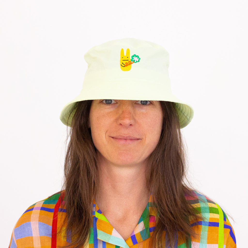 Smiling person wearing a purple bucket hat with a lime coloured bunny and green clover embroidery, paired with a colourful checkered shirt, standing in front of a yellow background with abstract orange shapes